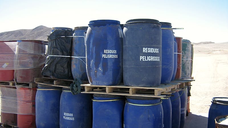 Rows of sealed steel drums on a concrete pad inside a warehouse, each with a printed label facing outward.
