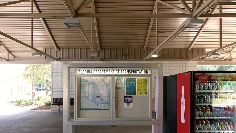 A cork noticeboard in a corridor with several printed sheets pinned in overlapping rows and a few loose pushpins.