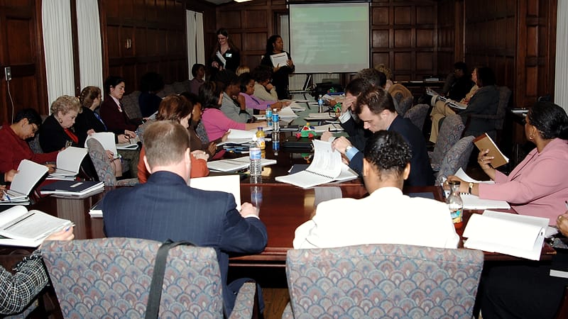 A person at a reception counter sliding a card across to a seated staff member who holds a pen over a clipboard.